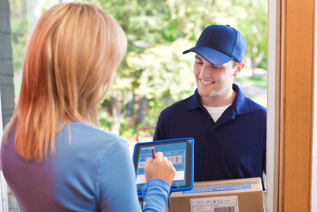 Delivery driver smiling as a customer signs for a package on a digital tablet at the doorstep.
